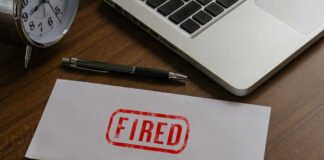 An envelope marked 'FIRED' on a wooden desk next to a clock and a laptop