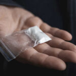 10 TONS Seized — Cartels Hammered Hand holding small bag of white powder.