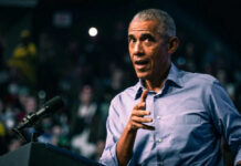 Barack Obama in blue shirt speaking at podium with microphone.