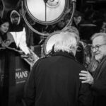 Two men conversing at a film premiere with a crowd in the background
