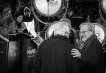 Two men conversing at a film premiere with a crowd in the background