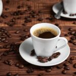 Coffee Apocalypse Brewing A white coffee cup filled with coffee on a wooden table surrounded by coffee beans