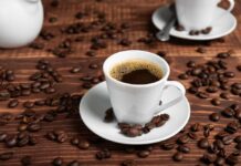 A white coffee cup filled with coffee on a wooden table surrounded by coffee beans