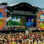 LEGENDARY Woodstock Icon DIES — Untold Military Past A large crowd at an outdoor music festival with a vibrant stage