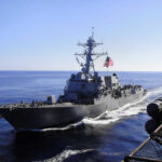 US Bombs Iran Amid Ceasefire Peace Talk Warship with American flag sailing in the ocean.